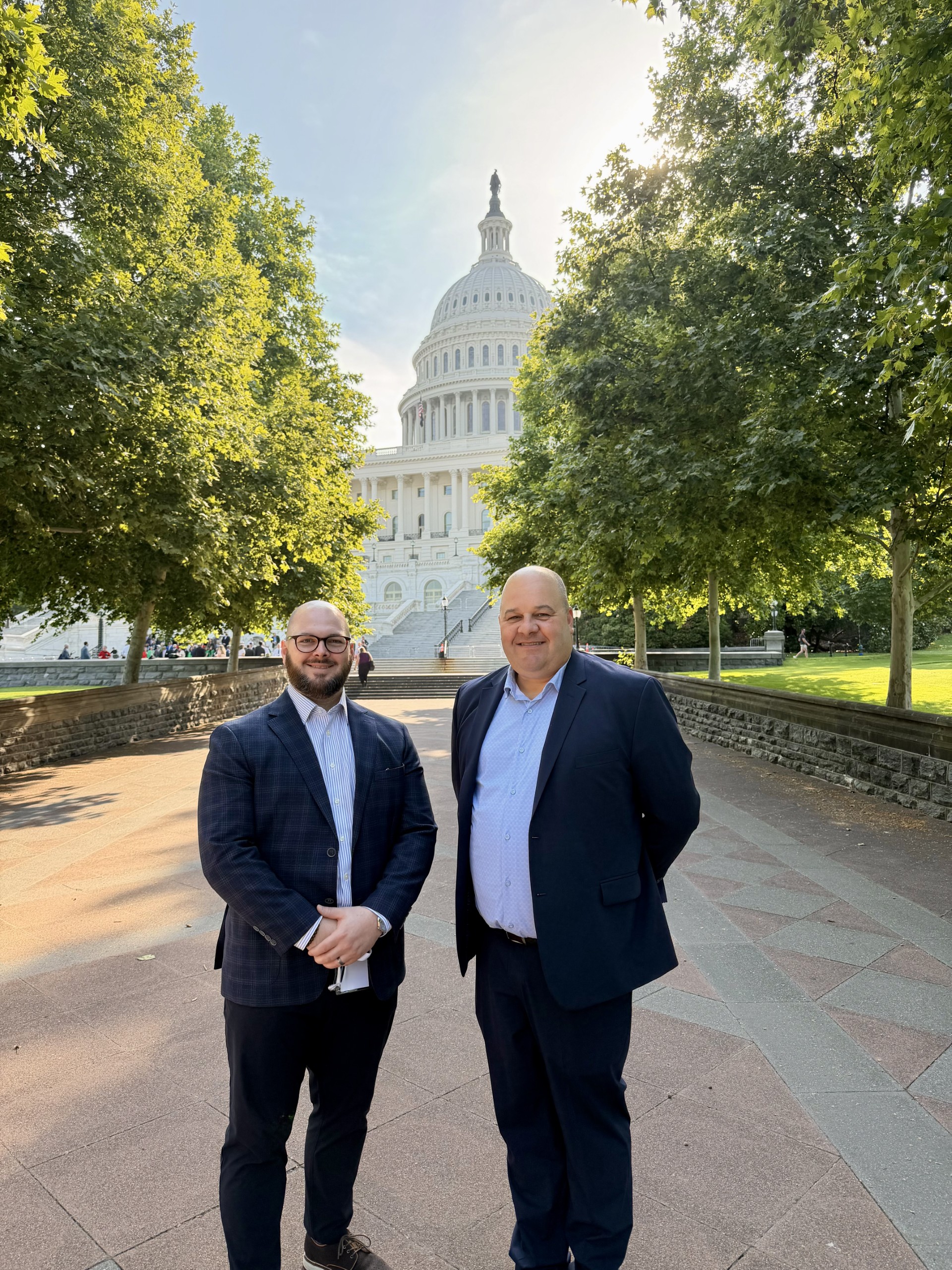 Two men in blazers stand on a walkway with the U.S. Capitol dome and trees behind them.