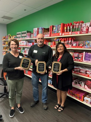A woman, a man and a woman each hold a framed plaque in a room lined with red candy displays.