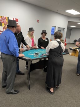 People, some in black hats, gather around a teal casino table with a dealer in a black apron in an office space.