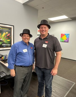Two men in black fedora hats pose together, one in a blue shirt and one in a gray polo.