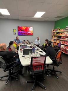 People seated around a white conference table with laptops, with a colorful screen on the wall and candy shelves at right.