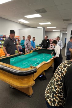 A craps table with teal felt and wooden rails, with players gathered at the far end of an office room.