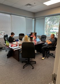 A few people work at desks pushed together in a corner room with window blinds.