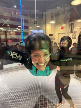 A smiling person in a helmet and goggles floats in an iFLY indoor skydiving tunnel.