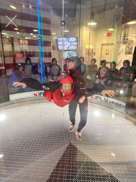 A person in a red flight suit and helmet floats in an indoor skydiving wind tunnel while onlookers watch.