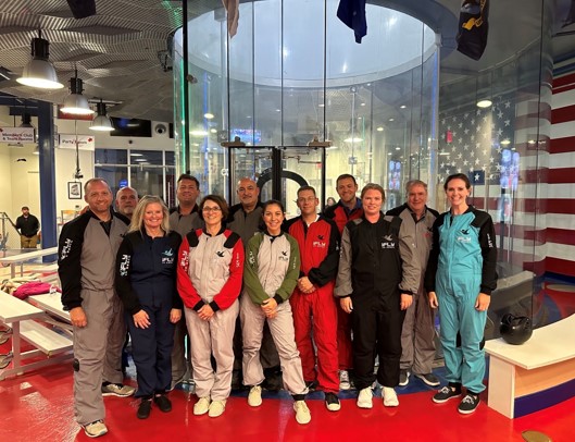 A group of adults in flight suits pose in front of an indoor skydiving wind tunnel.