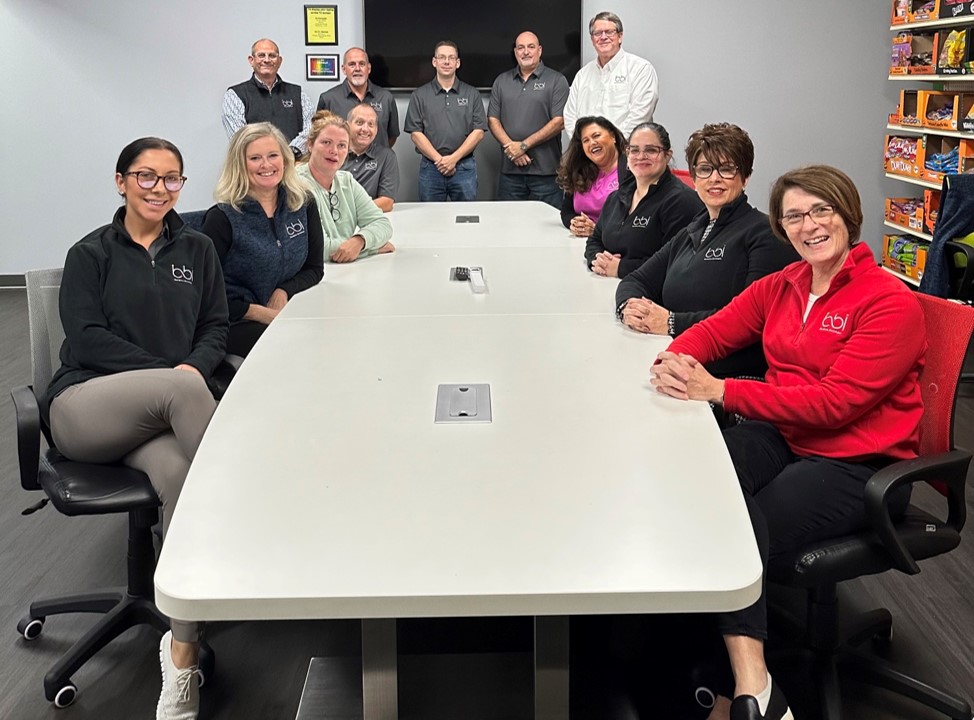 BBI staff in bbi-logo jackets and shirts sit and stand around a white conference table, with candy shelves behind.