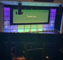 Stage seen from the audience, with two presenters and a screen reading Thank you between purple and green panels.
