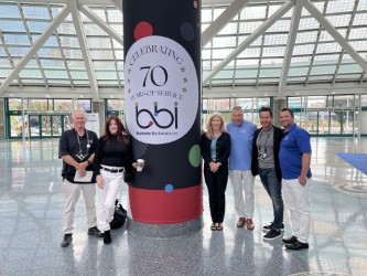 Six adults pose beside a column graphic reading Celebrating 70 Years of Service bbi in a glass-roofed convention hall.