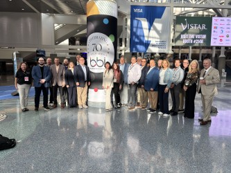 Large group of adults lines up in front of a bbi 70th anniversary column in a convention center concourse.