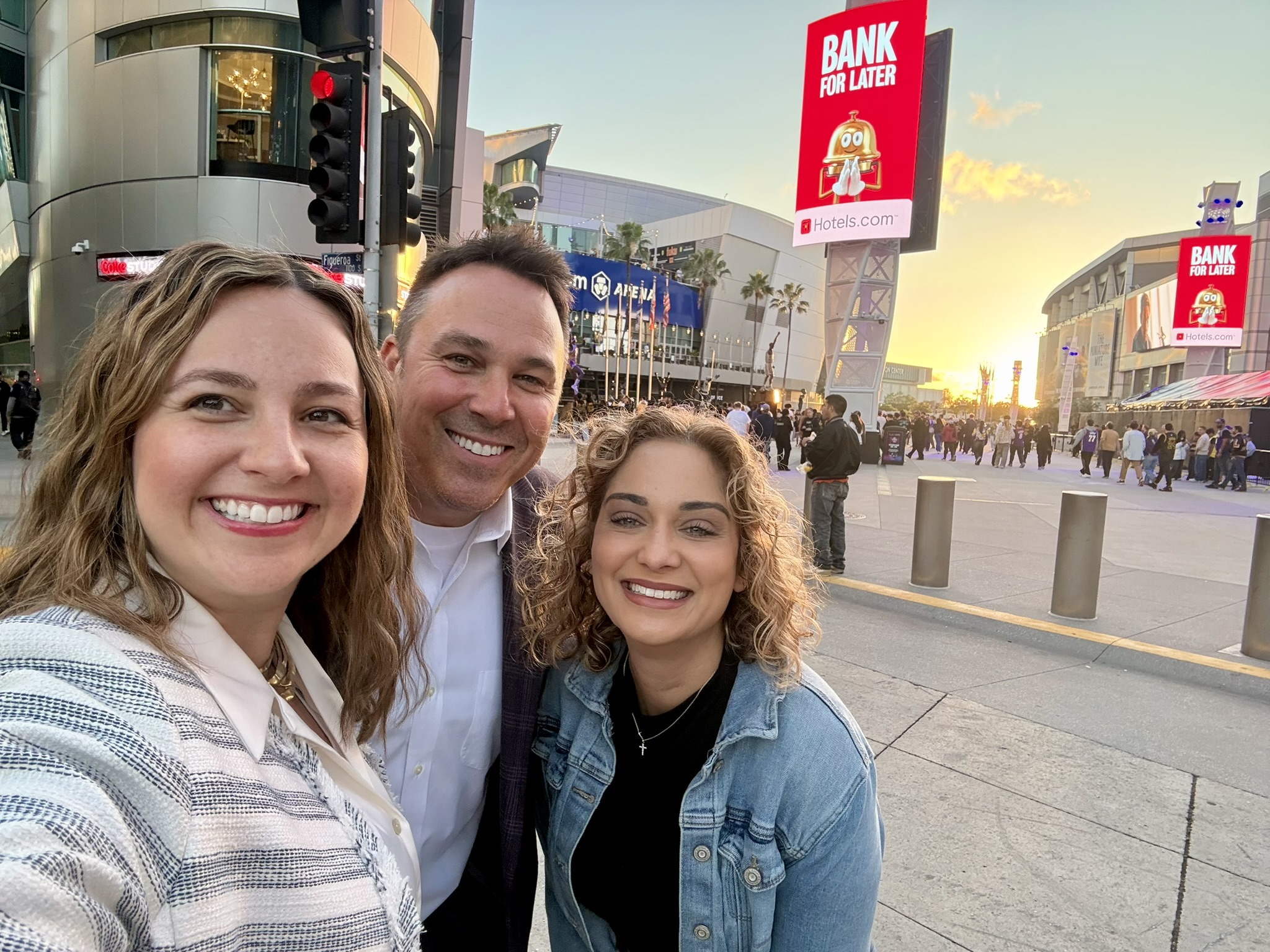 Three smiling people take a selfie outdoors at sunset, with arena buildings and Hotels.com billboards behind them.