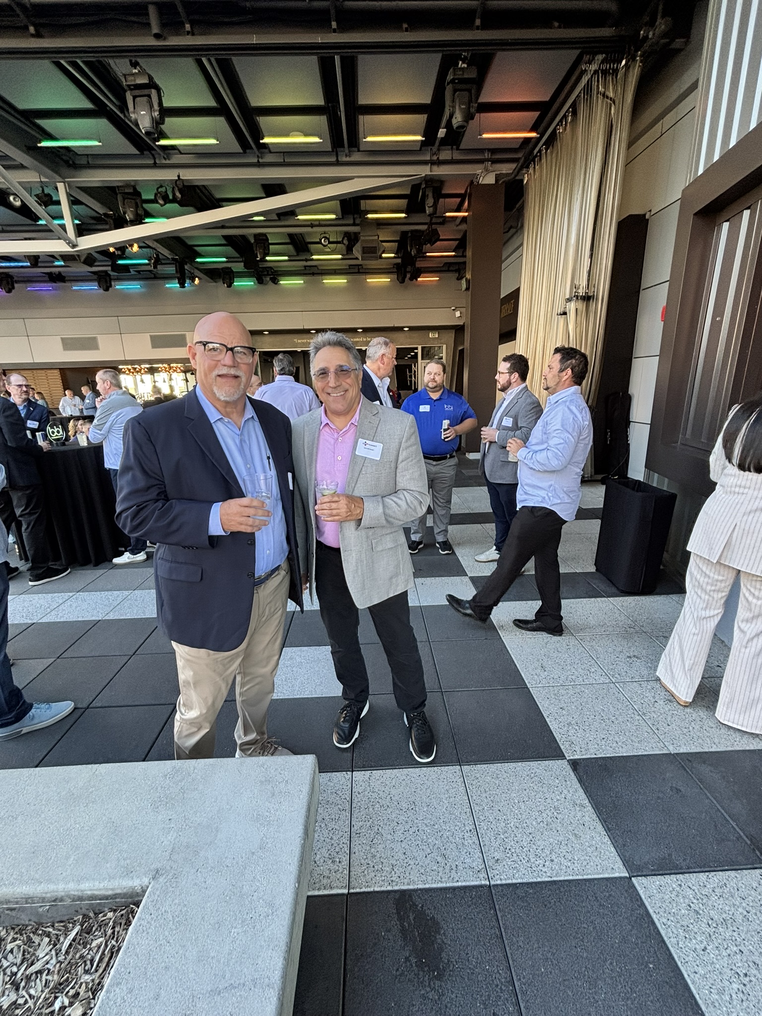 Two men in blazers hold drinks on a checkerboard-tiled terrace, with guests mingling behind them.