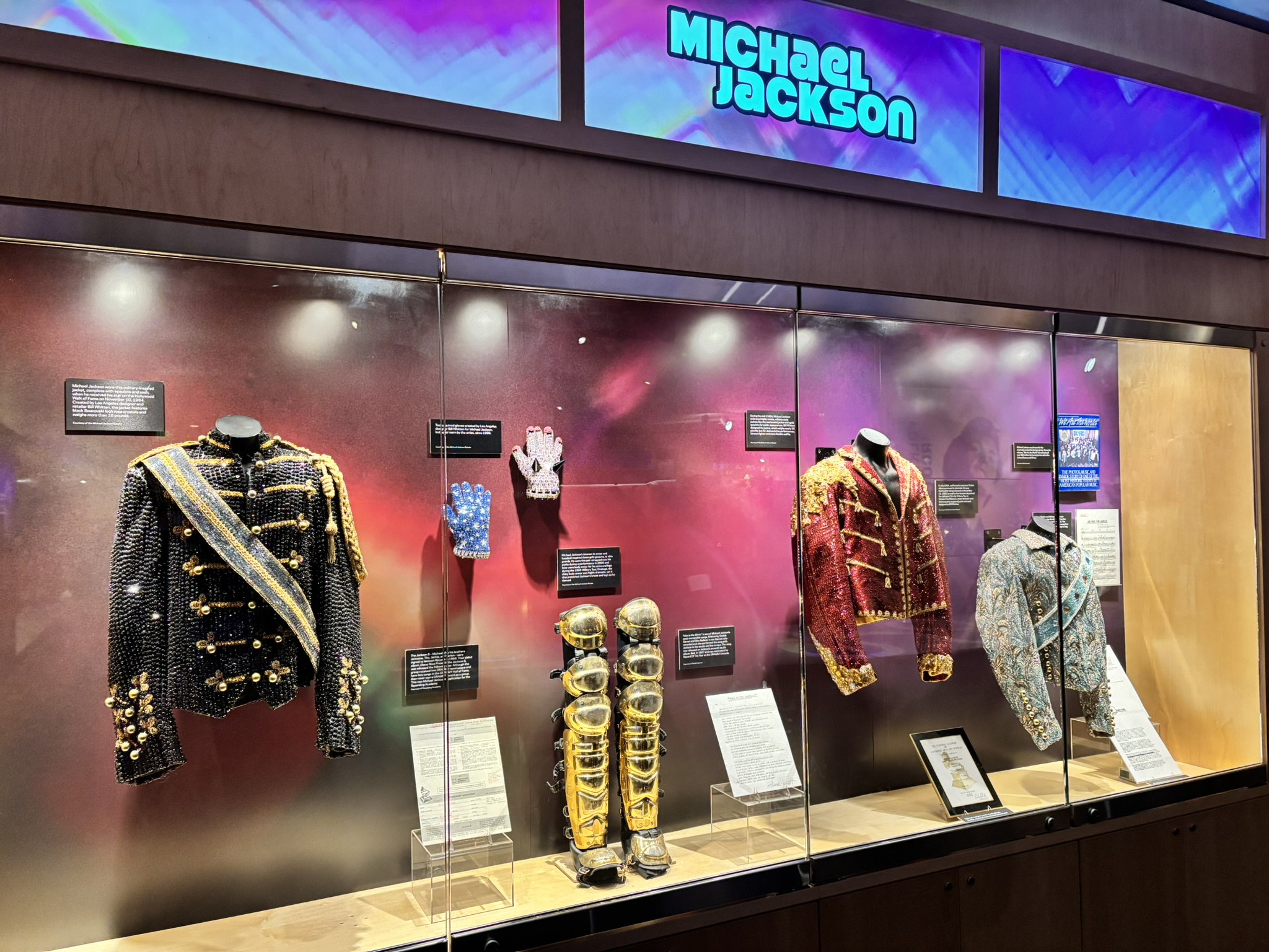 Museum display cases under a Michael Jackson sign holding sequined jackets, a crystal glove and gold leg guards.