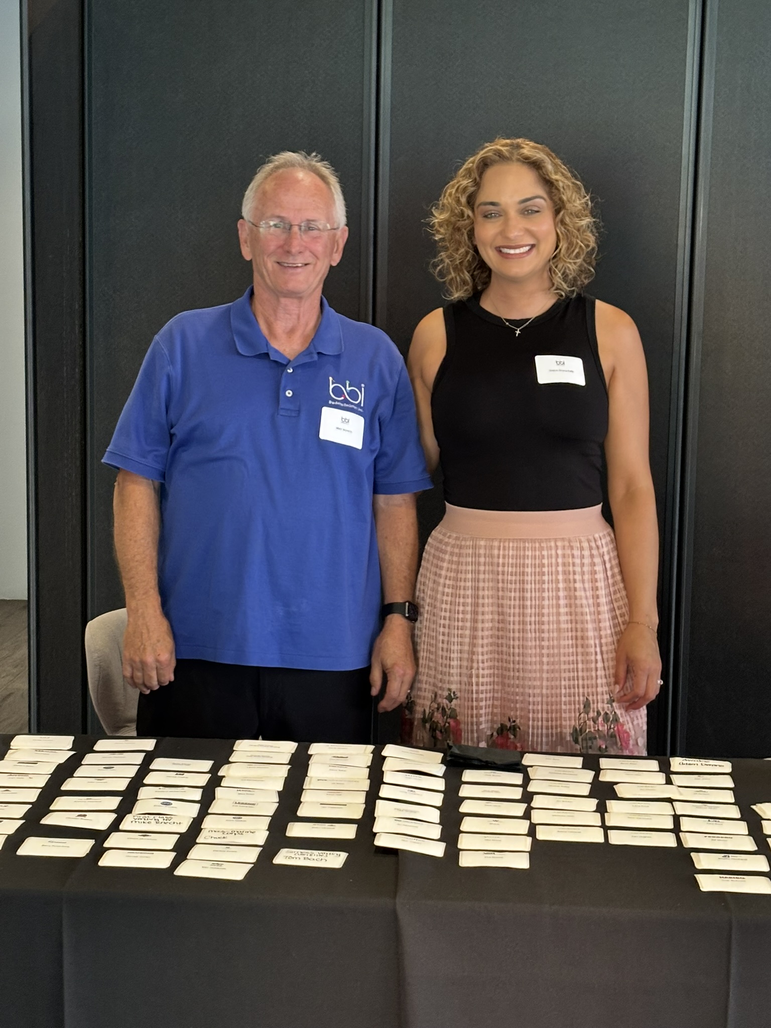 Man in a bbi polo and a woman stand behind a black table covered in handwritten name tags.