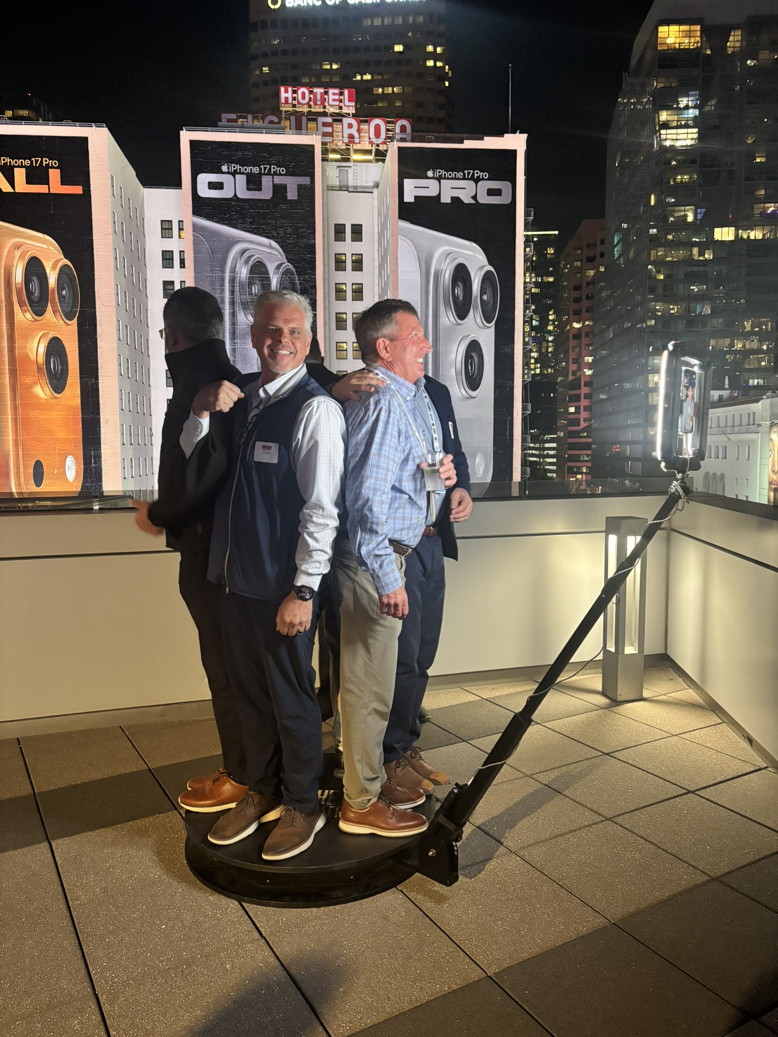 Three men pose back to back on a rooftop 360-degree photo platform, with lit city billboards behind.