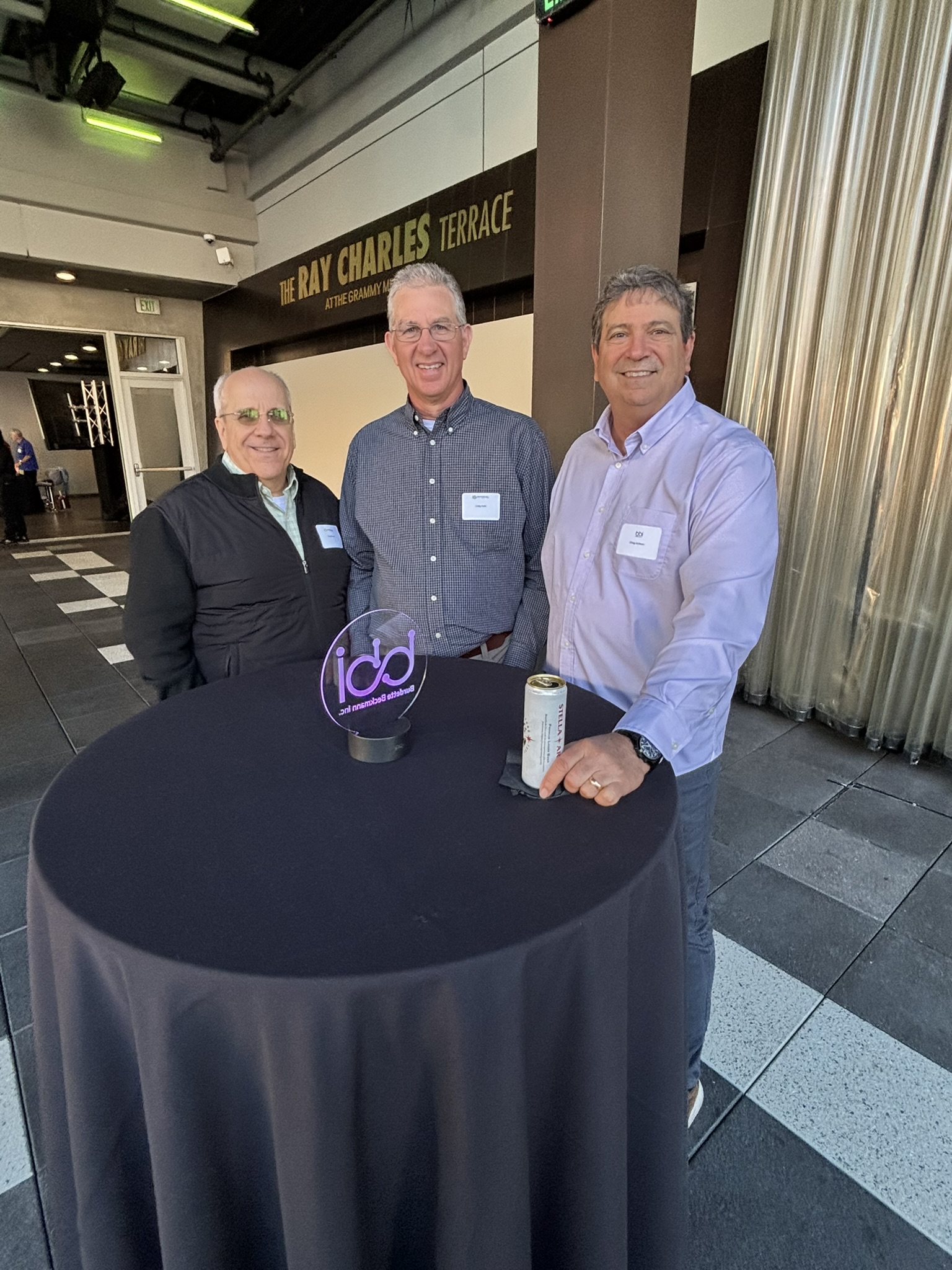 Three men stand around a draped cocktail table with a bbi glass logo, under a Ray Charles Terrace sign.