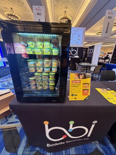 Lit glass refrigerator of Dole Whip cups on a table with a bbi table cover and a Dole Whip sign.
