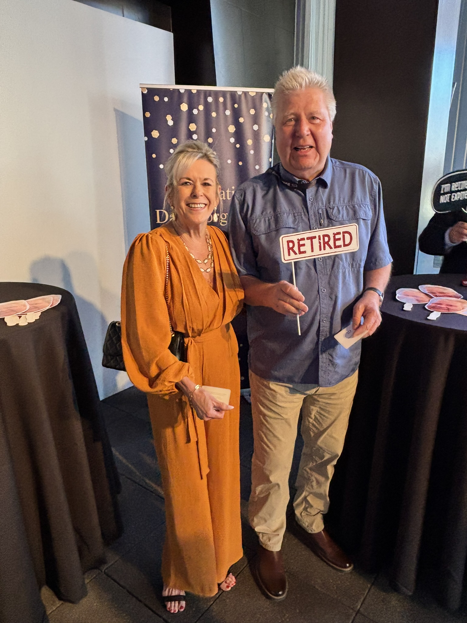 Man holding a Retired prop sign poses beside a woman in an orange jumpsuit at an evening reception.