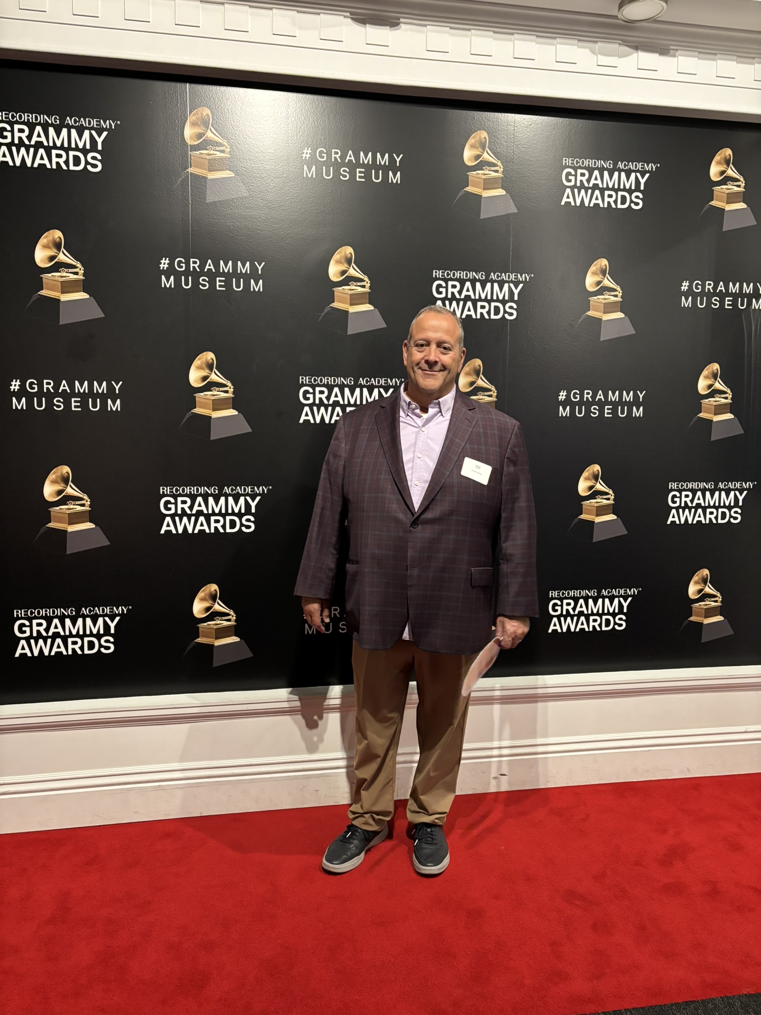 Man in a plaid blazer stands on a red carpet in front of a Grammy Awards and Grammy Museum backdrop.