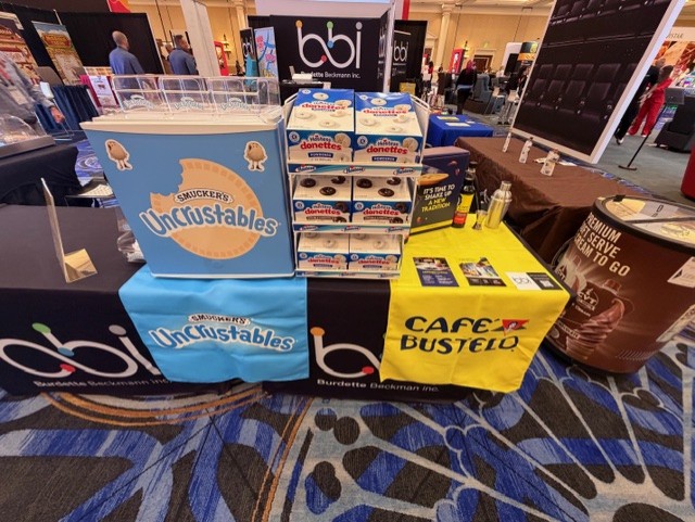 Smucker's Uncrustables, Hostess donettes and Cafe Bustelo displays on branded table covers beside a bbi table.
