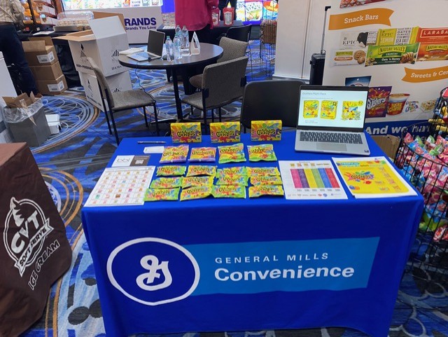 Blue General Mills Convenience table with Gushers fruit snack packs, a laptop and printed price sheets.