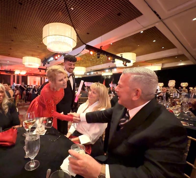 Woman in a red gown leans down to shake hands with a man in a dark suit seated at a gala table.