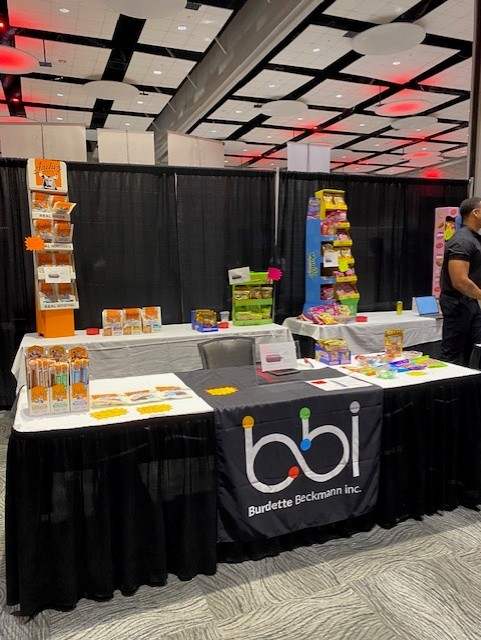 bbi booth table with orange and yellow snack racks and candy bags under a ballroom ceiling with red lights.