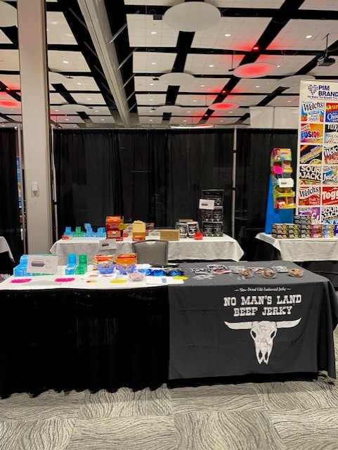 Table with a black No Man's Land Beef Jerky cover beside candy displays and a PIM Brands banner.
