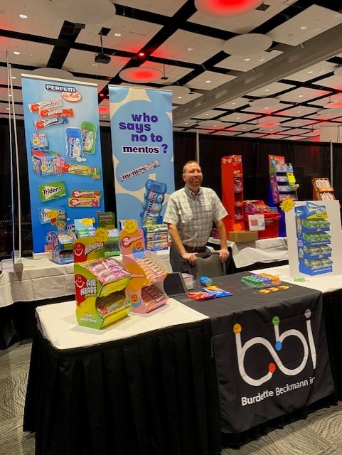 Smiling man stands behind a bbi table with Mentos and Perfetti Van Melle banners and Air Heads displays.