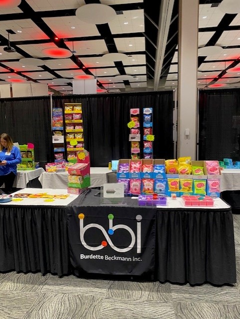 bbi booth table with boxes of gummy candy in pink, yellow and blue, and a woman in blue at the left.