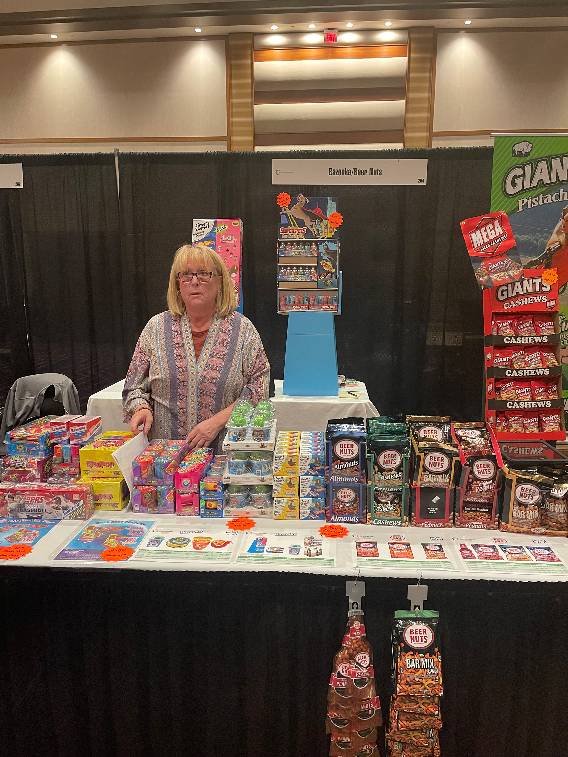 A woman in a patterned top stands behind a Bazooka and Beer Nuts table of candy and nut displays in a ballroom.