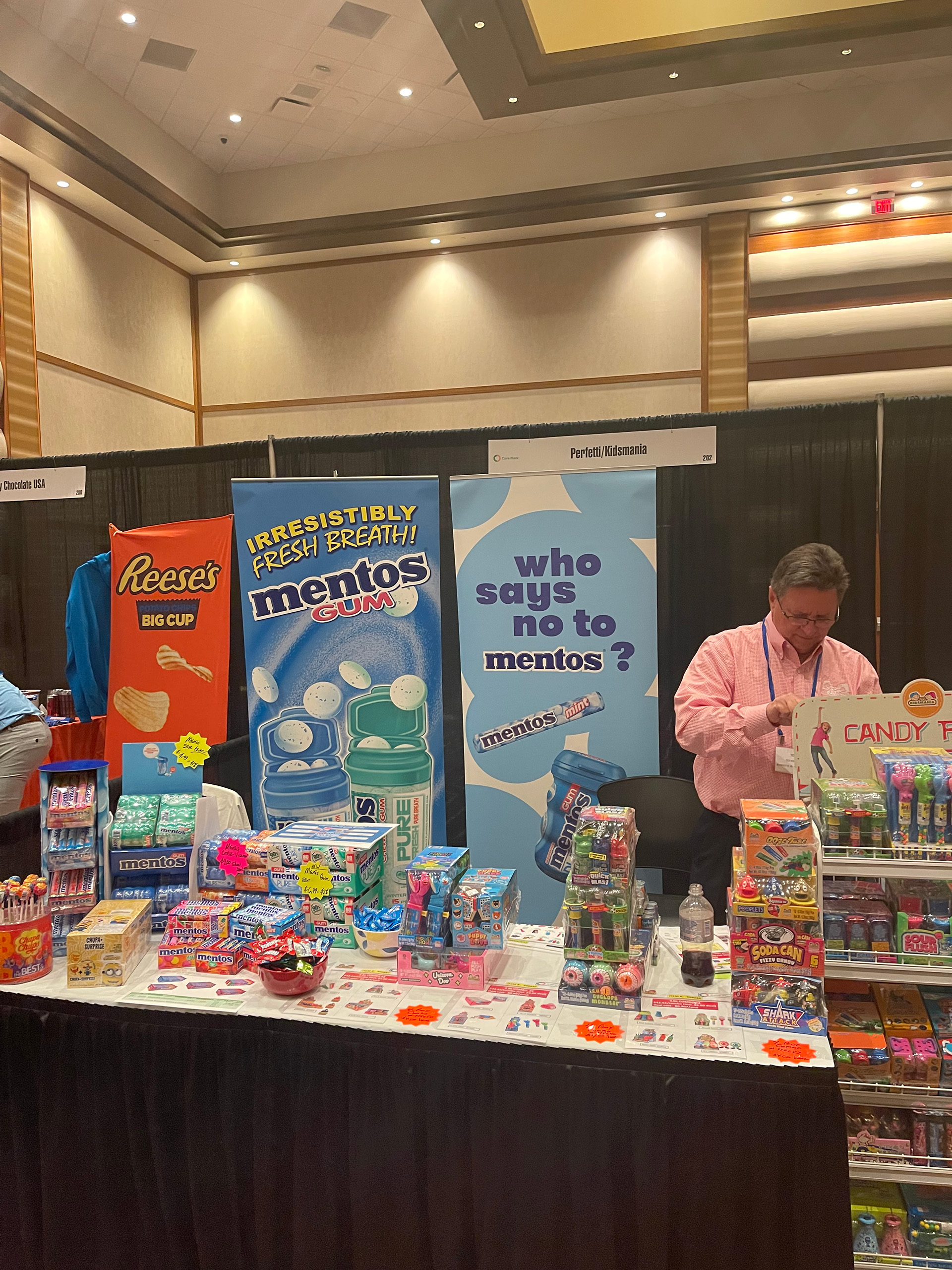 A man in a pink shirt stands behind a table of Mentos, PEZ and candy toys beneath two Mentos banners.
