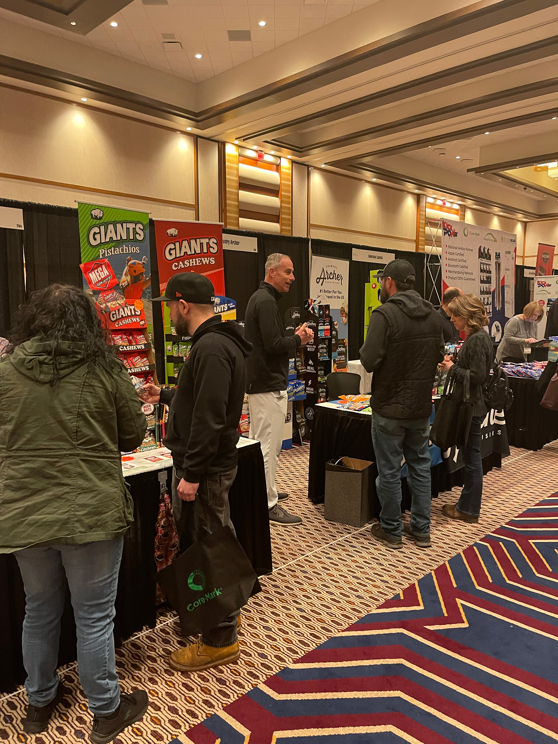 Attendees walk an aisle of black-draped tables with Giants pistachio and cashew banners, one carrying a CoreMark bag.