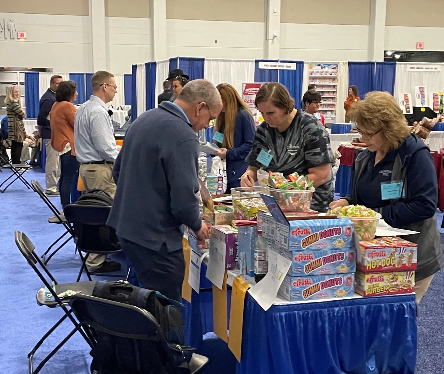 Attendees lean over a blue-skirted table of efrutti gummy donuts and candy at a trade show.