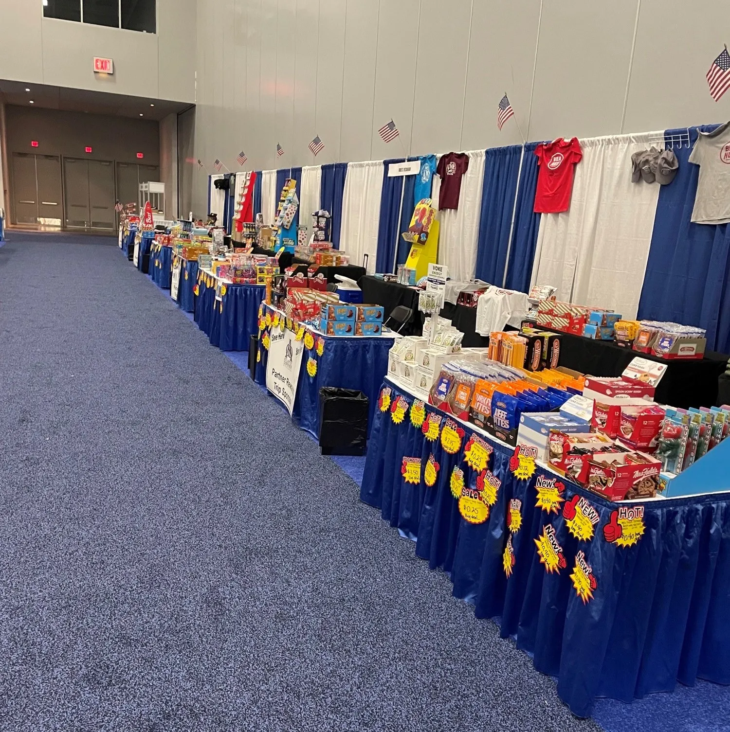 Long row of tables with blue skirts and snack and candy displays in a convention hall, with small American flags on the wall.