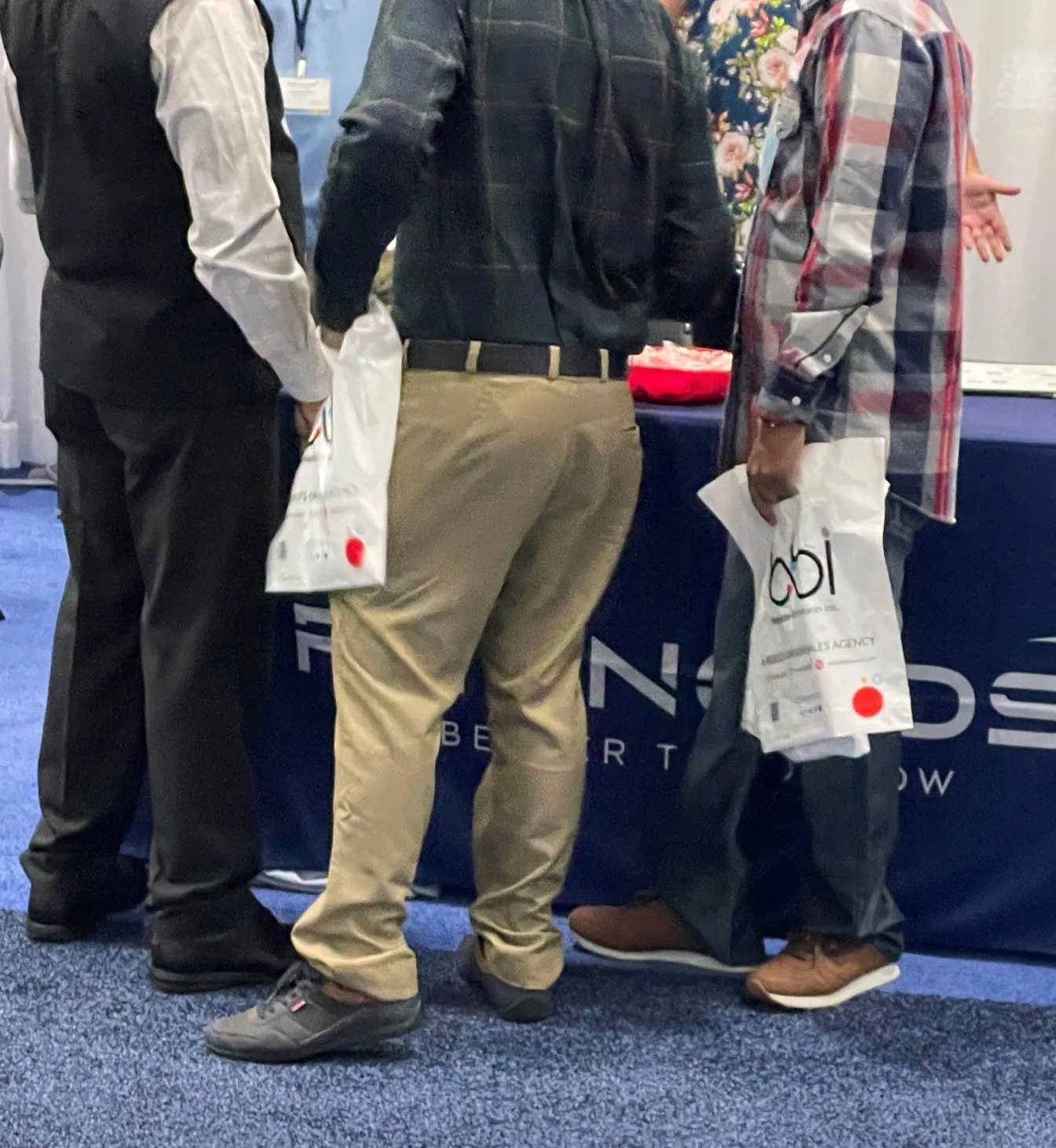 Attendees seen from behind at a navy-draped table, holding white shopping bags, one printed with the bbi logo.