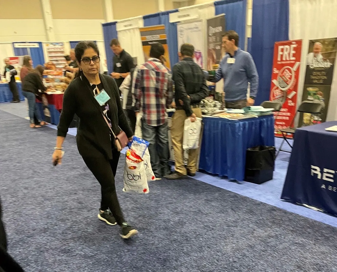 A woman in black walks an aisle carrying a white bbi bag, with attendees talking at booths behind her.