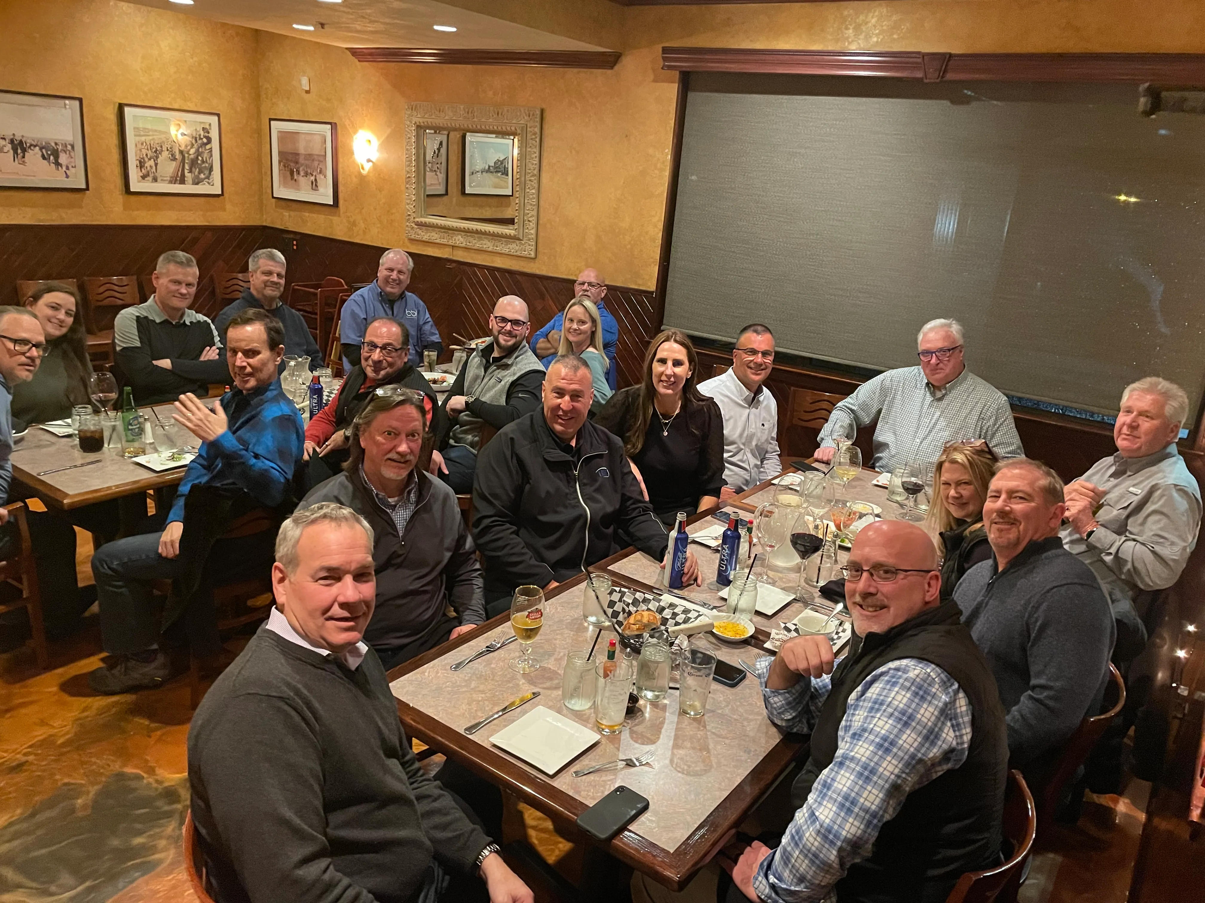 About twenty people seated at long restaurant tables with wine glasses, many smiling toward the camera.
