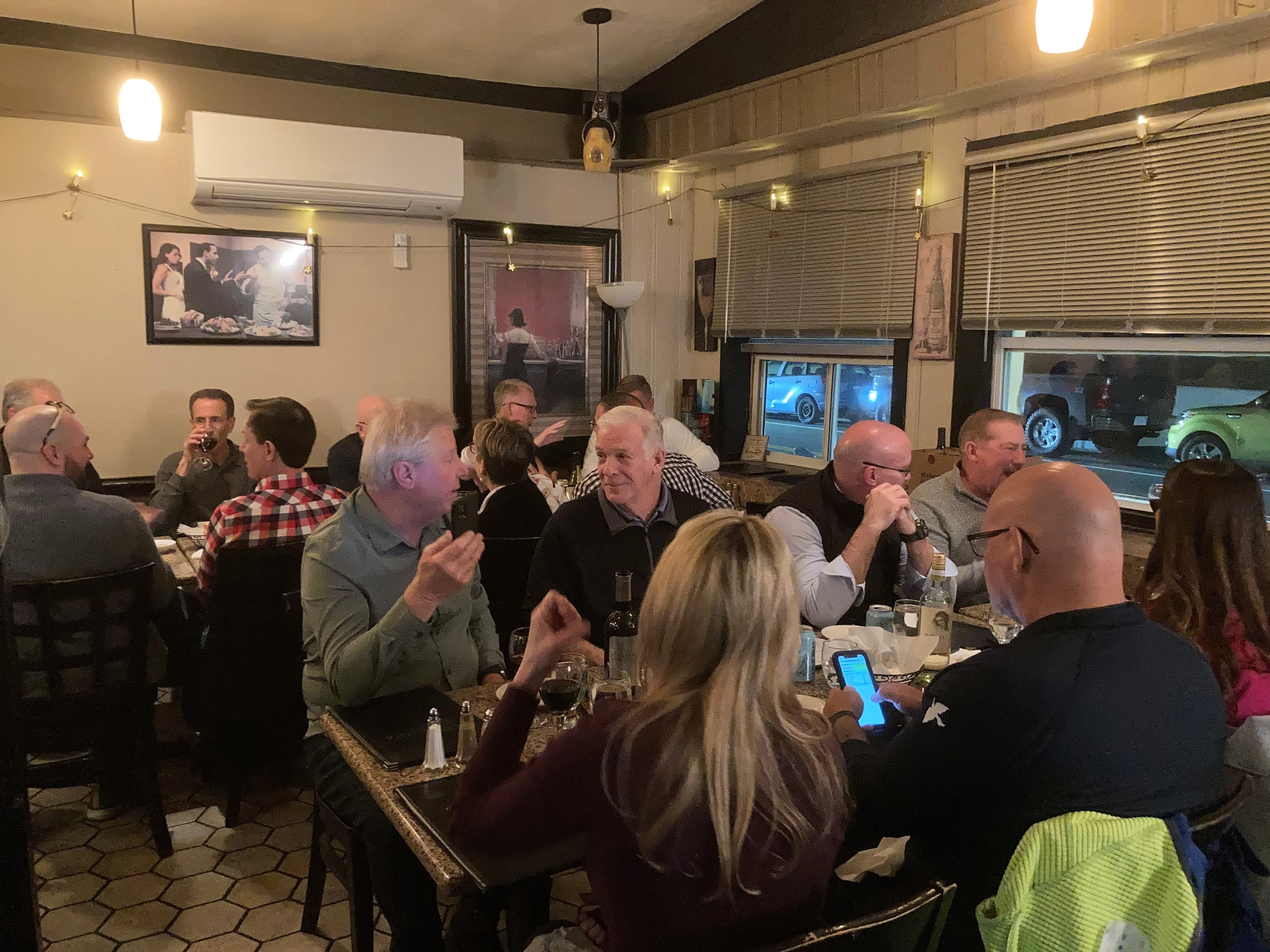 Diners talk at tables in a small restaurant, one man holding up his phone, with windows showing parked cars.