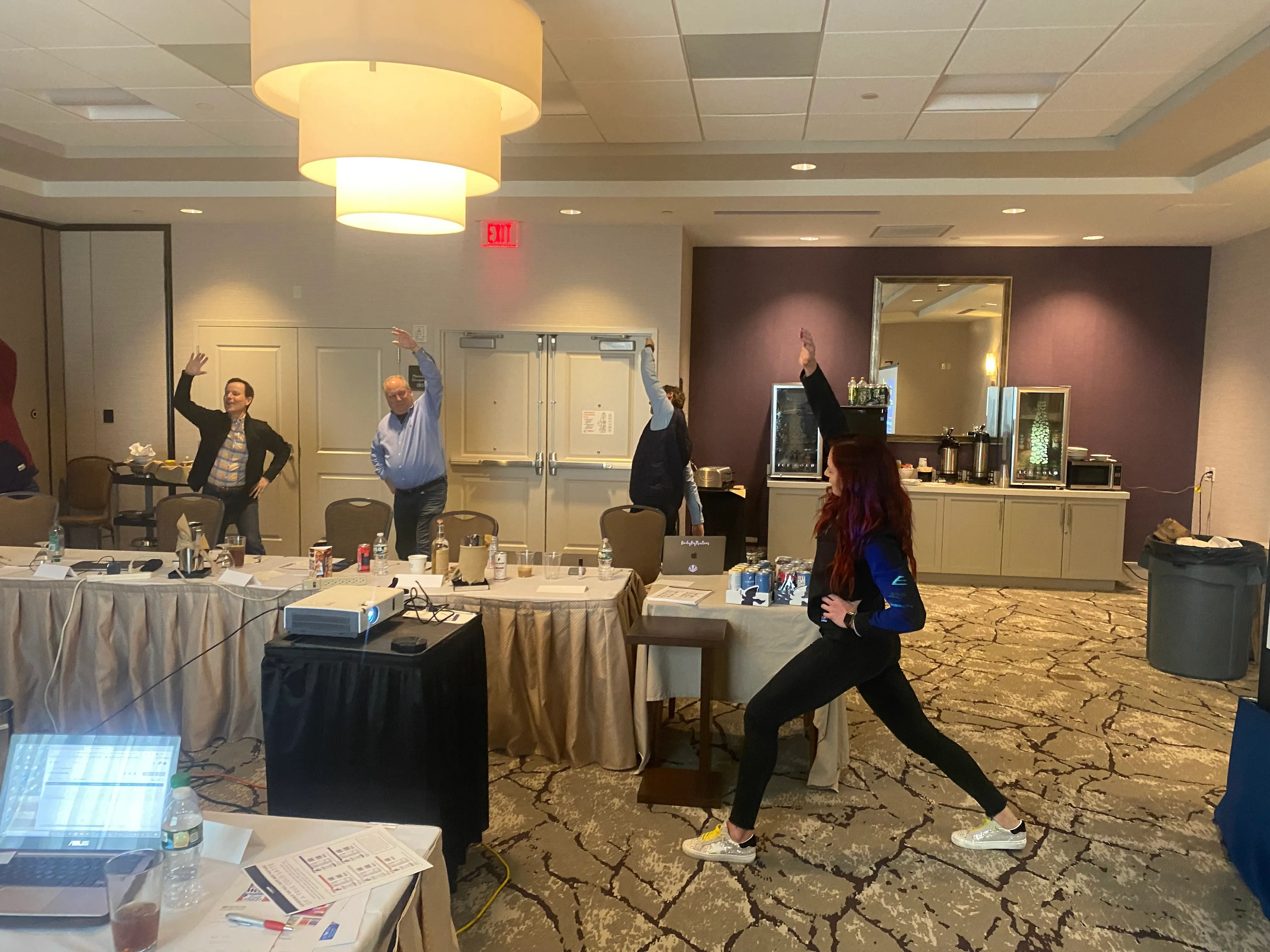 A woman lunges in a stretch while others reach overhead in a meeting room with a projector and coffee station.