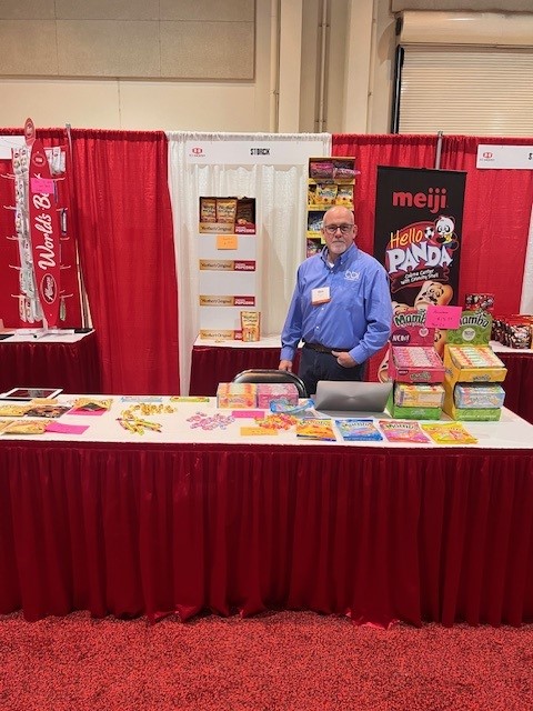 Man in a blue shirt at a red-skirted booth table with candy samples, near a Meiji Hello Panda banner.