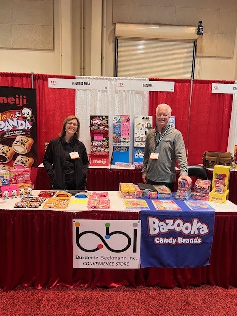 Woman and man at a booth with a bbi Convenience Store sign, a Bazooka Candy Brands table runner and a Meiji banner.