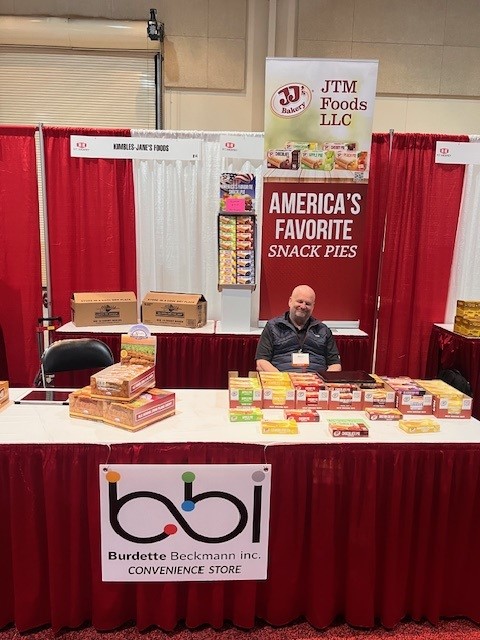 Man seated behind a bbi Convenience Store table under a JTM Foods banner reading America's Favorite Snack Pies.