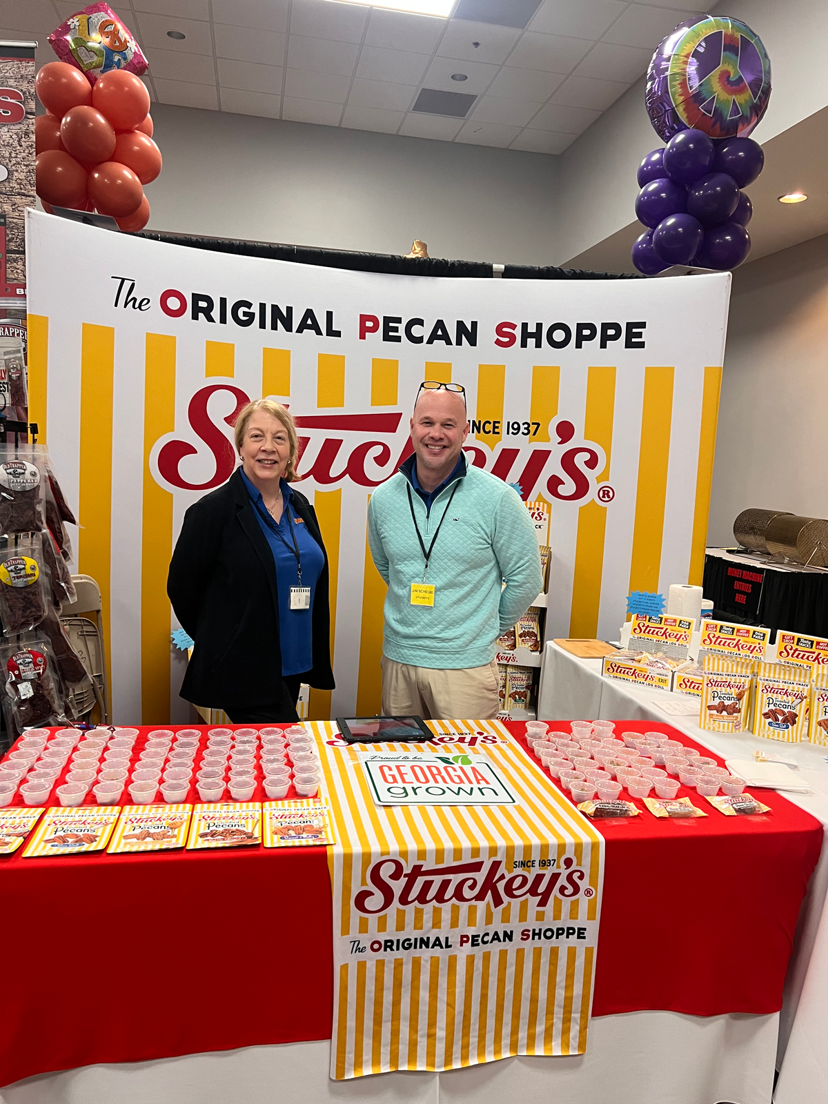 Woman and man with badges stand at a Stuckey's Original Pecan Shoppe booth with a red table and rows of sample cups.