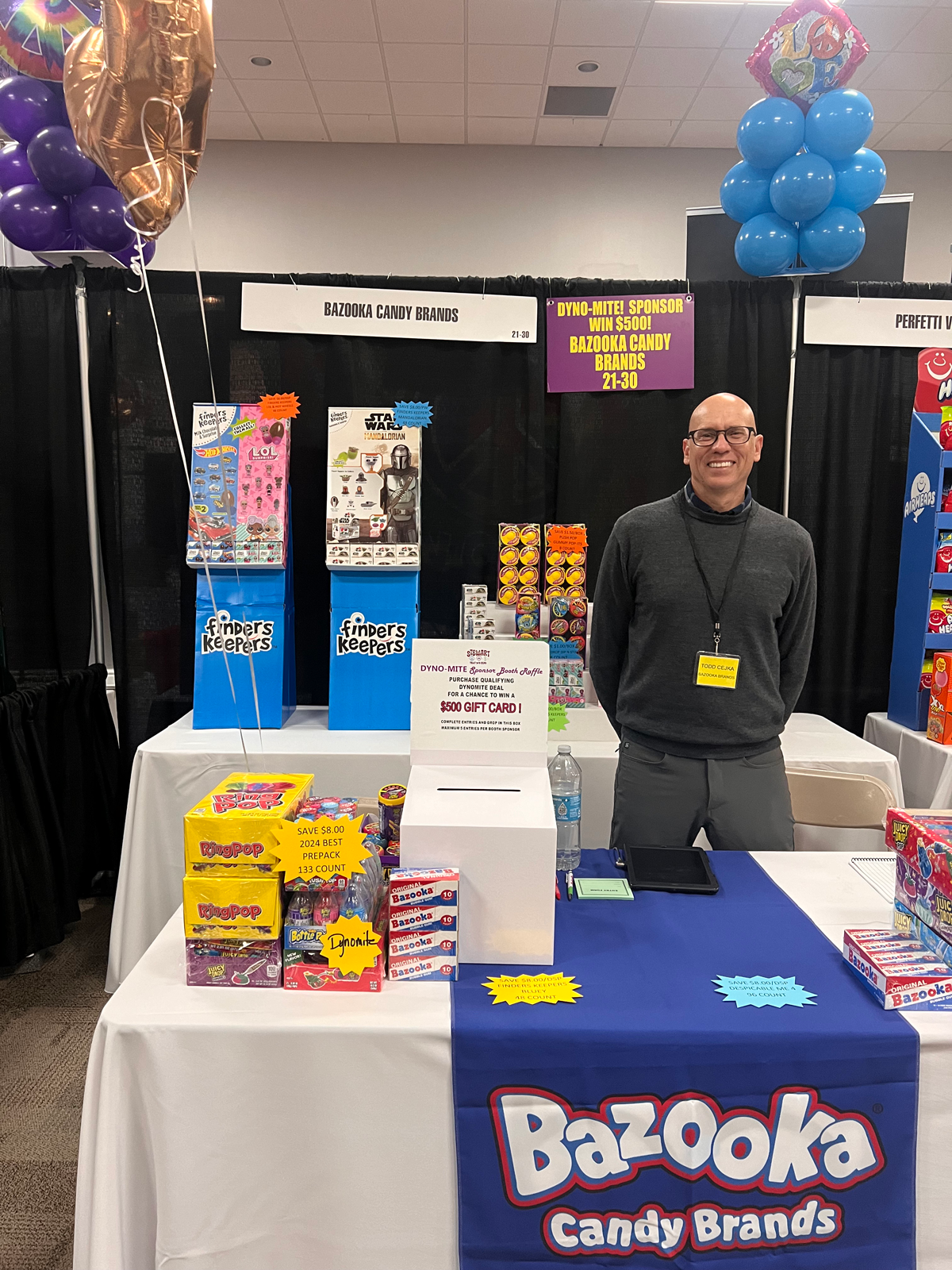 Man in a gray sweater at a Bazooka Candy Brands booth with a blue Bazooka table runner and Finders Keepers displays.