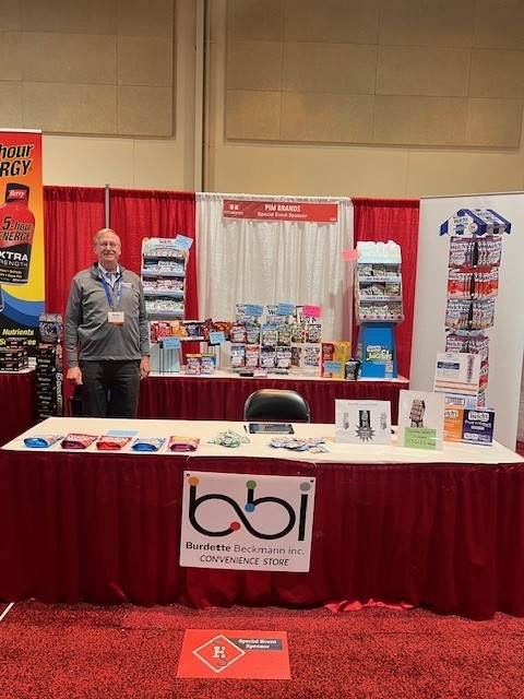 Man in a gray quarter-zip at a bbi Convenience Store table with a PIM Brands sign and a 5-hour ENERGY banner at left.