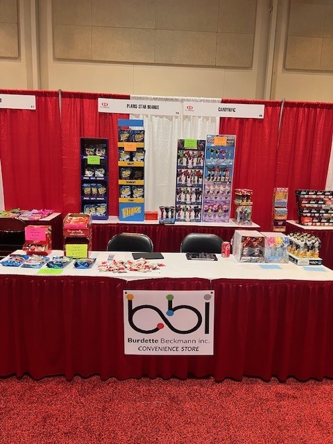Unstaffed booth with a bbi Convenience Store sign, Pez and snack racks, and two black chairs.