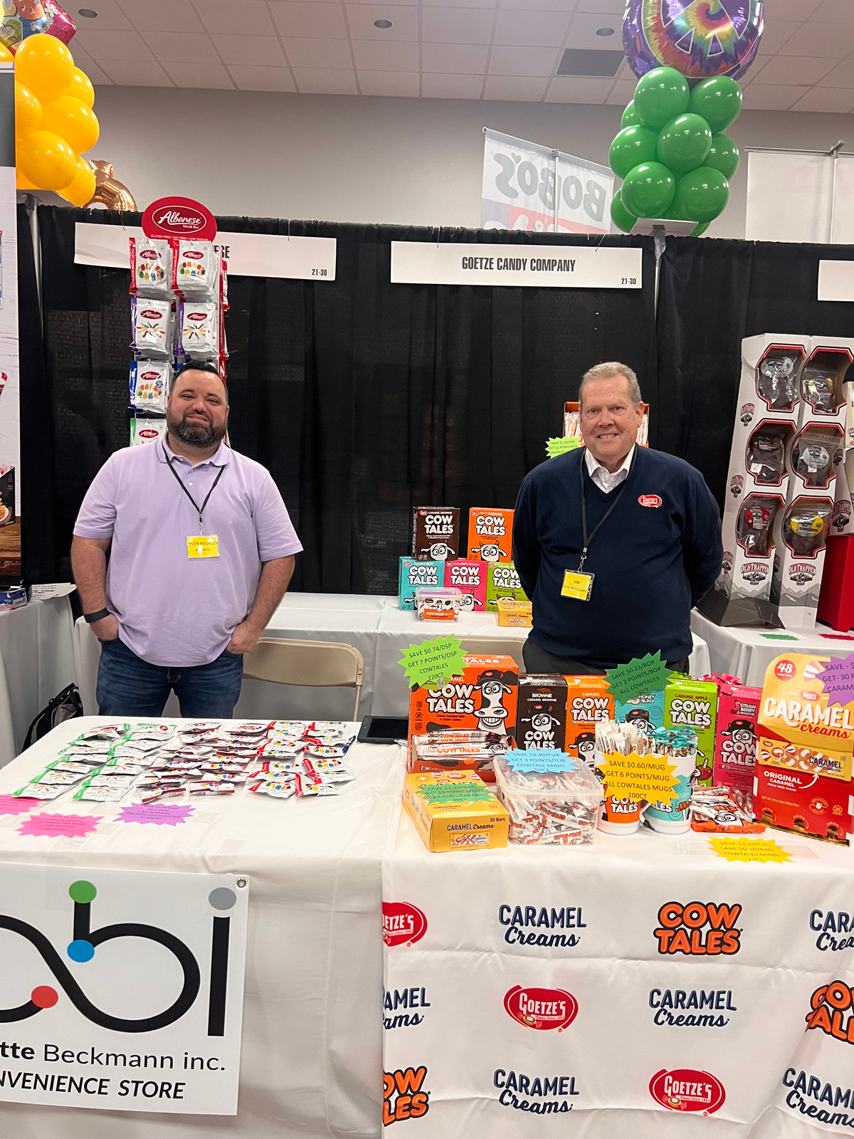 Two men at a Goetze Candy Company booth with a Cow Tales and Caramel Creams tablecloth and a bbi Convenience Store sign.