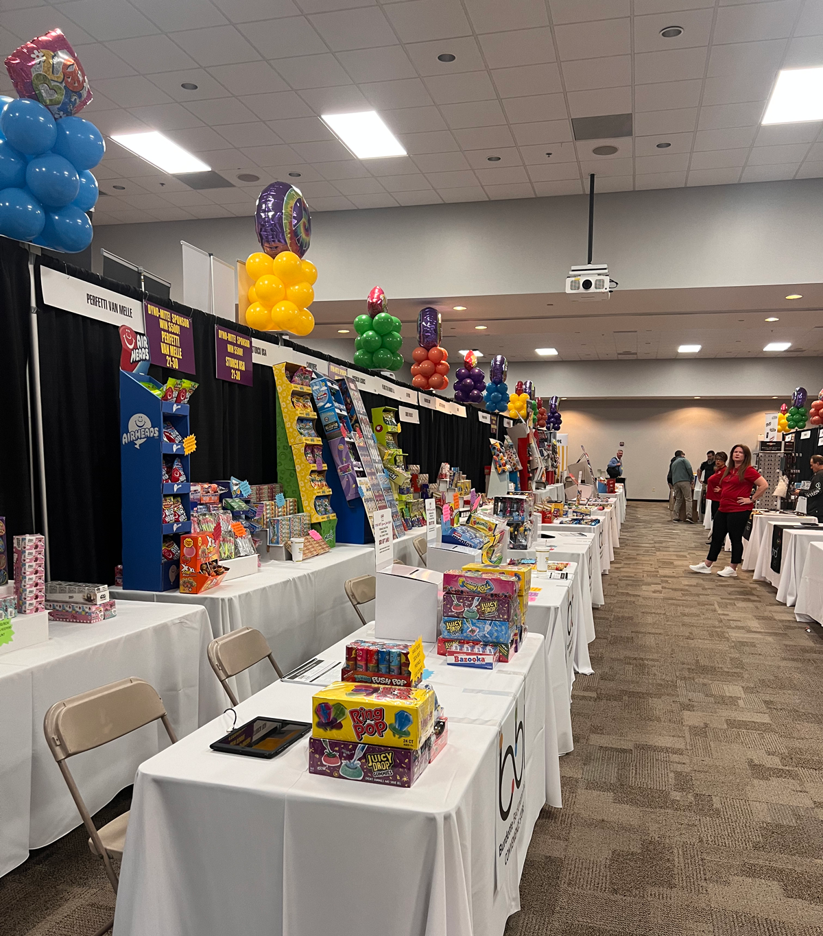 Row of white-draped trade show tables with candy displays and balloon columns stretching down a hall.