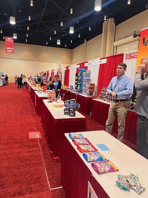 Aisle of red-skirted booths in a convention hall under an Aisle 800 sign, with two men standing at right.